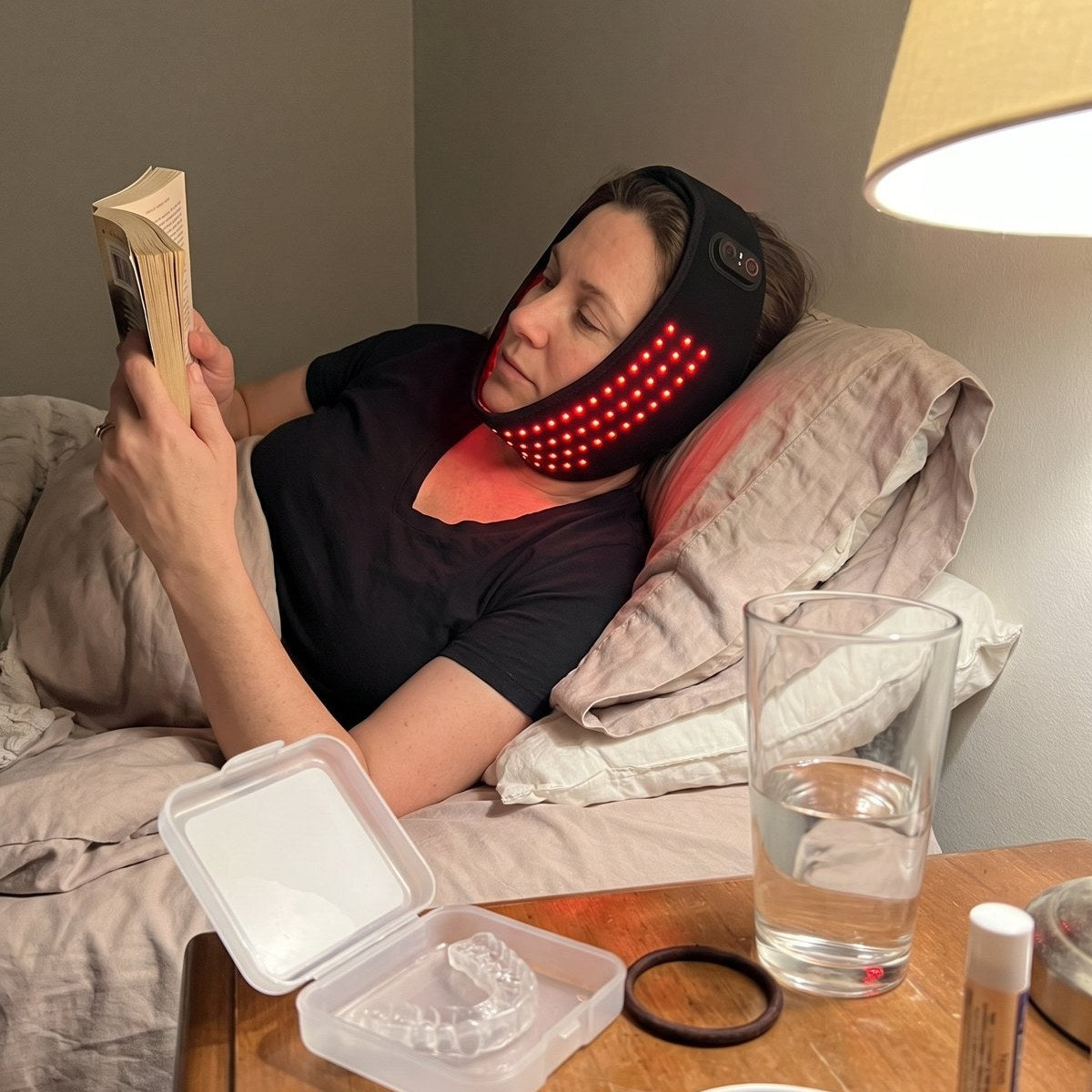 A phone photo of a cluttered nightstand, a woman reading in bed behind it with the jaw pad on.